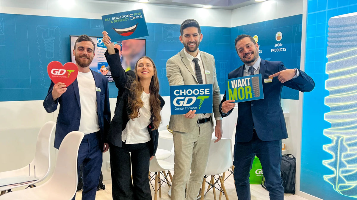GDT team members stand together in a bright blue booth, holding signs including a red heart-shaped 'GDT' sign, a blue 'CHOOSE GDT' sign, and a blue 'WANT MOR' sign.