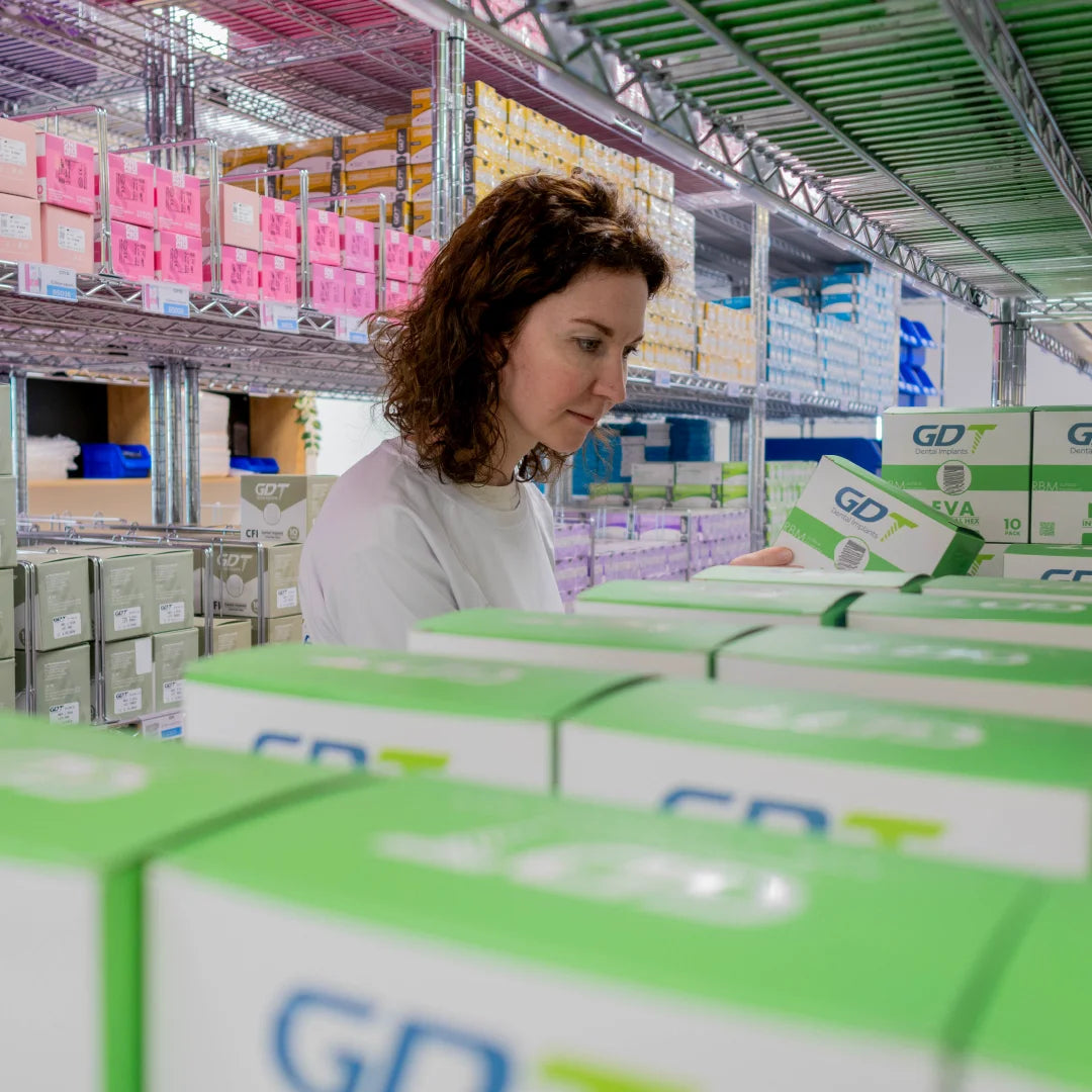 warehouse worker, holding a white and green GDT box while shelves of similar boxes line both sides.