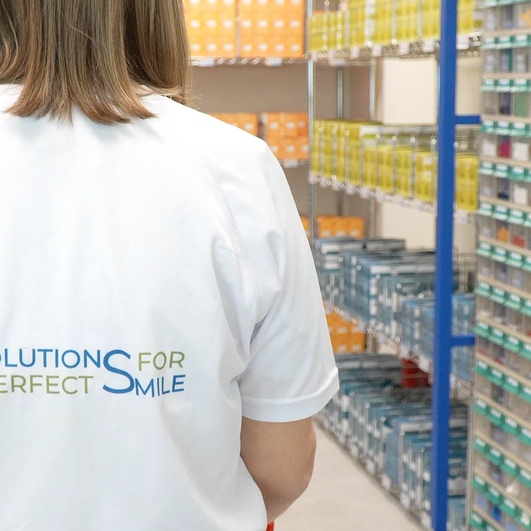 A person viewed from behind wearing a white short-sleeve T-shirt with the text 'ALL SOLUTIONS FOR PERFECT SMILE' printed on the back in blue and olive green, standing in a retail aisle with shelves of packaged products in the background.